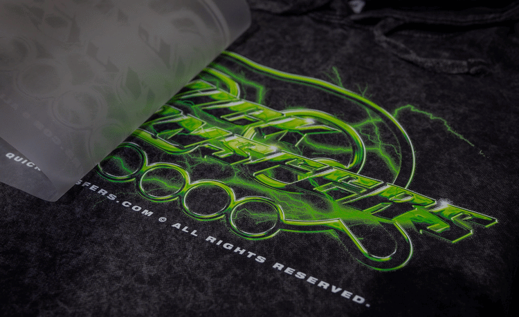 Close-up of a vibrant, lightning-themed graphic design in green and black on a dark fabric. Part of the design is covered by a translucent sheet. Text reads: