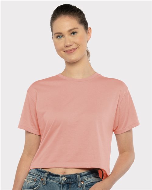 Load image into Gallery viewer, A person wearing a Next Level Women's Ideal Crop Top in peach made from combed ring-spun cotton and blue jeans stands before a plain light gray backdrop, softly smiling.