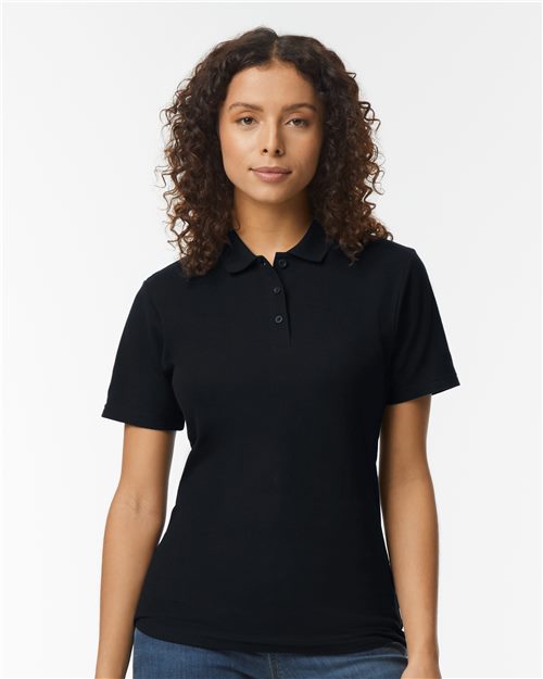 Load image into Gallery viewer, A woman with curly hair wears a Gildan Softstyle® Women's Pique Polo in black, made from OEKO-TEX certified ring-spun cotton. She stands against a white background, gazing directly at the camera, embodying sustainable manufacturing.