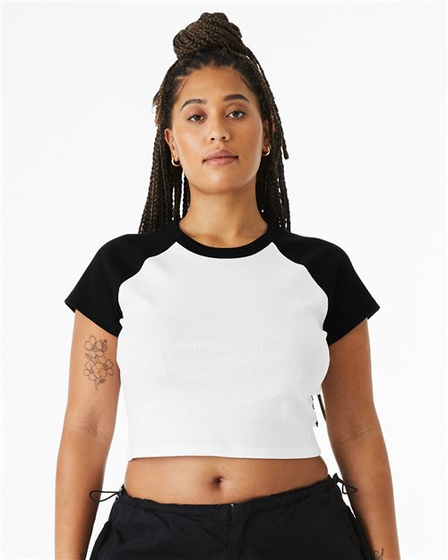 Load image into Gallery viewer, A person with long braids wears a black and white BELLA + CANVAS Women's Micro Rib Raglan Baby Tee made from Airlume combed cotton, paired with black pants. They have a tattoo on their left arm and stand against a white background, promoting sustainable manufacturing practices.