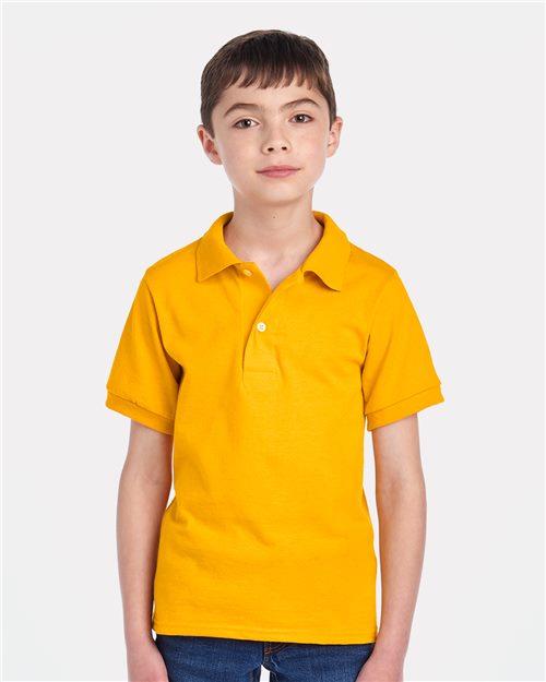 Load image into Gallery viewer, A young boy stands in a JERZEES - Youth Dri-Power® Polo and blue jeans against a plain white background, looking directly at the camera with a neutral expression.