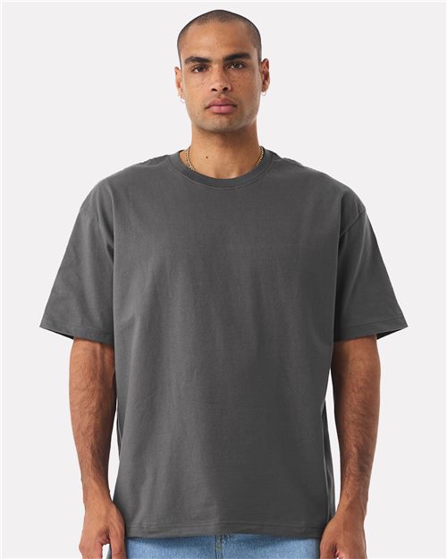 Load image into Gallery viewer, A person wears a BELLA + CANVAS - 7.5 oz Heavyweight Tee, showcasing its simple gray design with a unisex silhouette, paired with light blue jeans. Standing against a white backdrop, the individual has short hair and facial hair, maintaining a neutral expression and relaxed posture with hands at their sides.