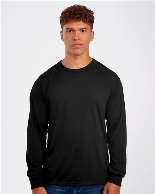 Load image into Gallery viewer, A young man with curly hair wears a plain black JERZEES - Dri-Power® Performance Long Sleeve T-Shirt made of moisture-management polyester and blue jeans, standing against a light gray background.