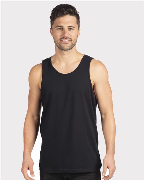 A person with short dark hair is smiling at the camera while wearing a Next Level - Cotton Muscle Tank, a sleeveless black top made from combed ring-spun cotton. The background is plain light gray, highlighting the brand's socially conscious manufacturing ethos.