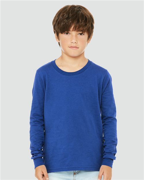 Load image into Gallery viewer, A young boy with brown hair stands against a light gray background, wearing a BELLA + CANVAS Youth Jersey Long Sleeve Tee in blue, made from Airlume combed cotton, paired with light blue jeans. He has a neutral expression, hands by his sides, showcasing the unisex sizing and sustainable manufacturing of the brand.