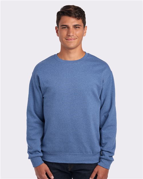Load image into Gallery viewer, A person in a blue JERZEES - NuBlend® Crewneck Sweatshirt and dark jeans stands against a plain white background, smiling at the camera.