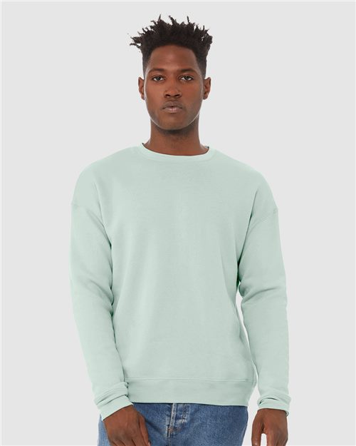 Load image into Gallery viewer, Wearing a light mint green BELLA + CANVAS Sponge Fleece Drop Shoulder Crewneck Sweatshirt made from Airlume combed and ring-spun cotton, a person pairs it with blue jeans against a plain gray background, exuding confidence and embodying sustainable manufacturing while looking directly at the camera.