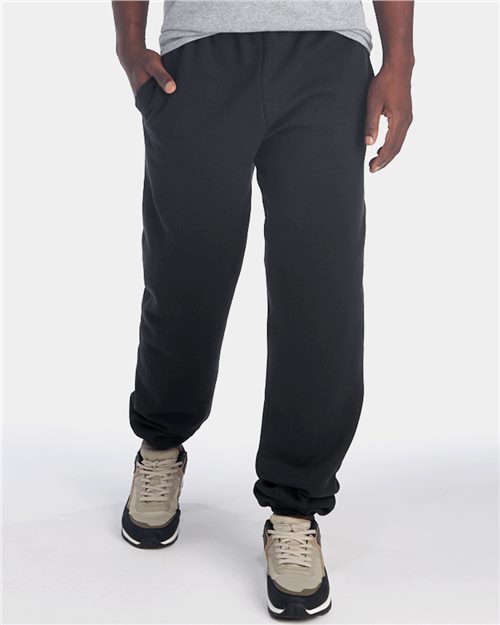 Load image into Gallery viewer, A person against a white background wears JERZEES Super Sweats NuBlend® Sweatpants with Pockets in black and gray sneakers, casually resting one hand in their pocket.