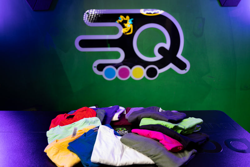 Load image into Gallery viewer, A stack of vibrant Quick Transfers Short Sleeve Shirt Mystery Bundle tees sits on a dark table before a green wall with their logo—perfect for spring cleaning sales or adding surprise variety to your wardrobe!.