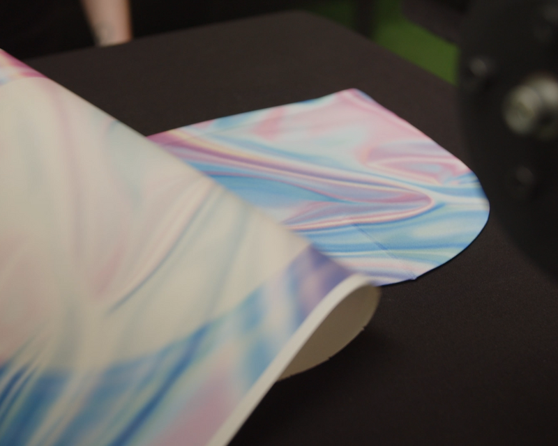 Load image into Gallery viewer, A close-up of a partly unrolled fabric roll with a soft, swirled pastel pattern in blue, pink, and white lies on a black surface. The silky texture's vibrant design is ideal for sublimation printing and bulk orders. Product: SUBLIMATION TRANSFERS - Transfers By Size by Quick Transfers.