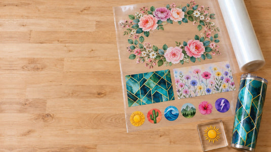 Wide UV DTF gang sheet on a wood workbench holding a floral design, two tumbler wraps and a row of round stickers, next to a wrapped tumbler and a clear acrylic block with a sun sticker