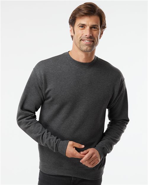 A man with short brown hair is wearing a black Hanes Perfect Fleece Crewneck Sweatshirt and dark pants. He's standing, smiling, hands together against a plain white background, exuding simplicity and sustainability.