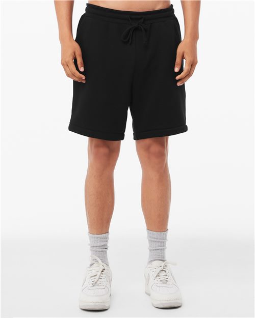 A person is standing against a plain white background, showing only the lower half of their body. They are wearing BELLA + CANVAS versatile black drawstring Sponge Fleece Shorts made from Airlume cotton, paired with gray socks and white sneakers.