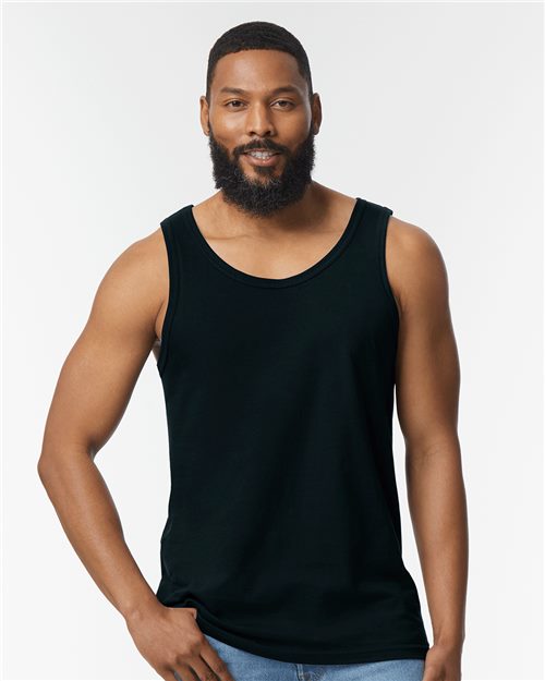 A bearded man in a Gildan - Softstyle® Tank Top, crafted from ring-spun cotton, stands against a white backdrop. With his right hand on his hip and a slight smile, he exudes relaxed confidence in socially conscious choices.