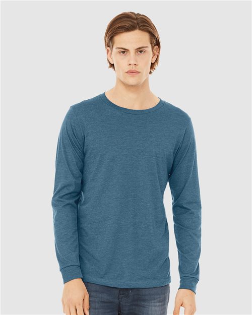 A person with medium-length hair wears a BELLA + CANVAS Heather CVC Long Sleeve Tee in teal and jeans, against a plain light gray background. They look directly at the camera, embodying sustainable manufacturing and unisex sizing.