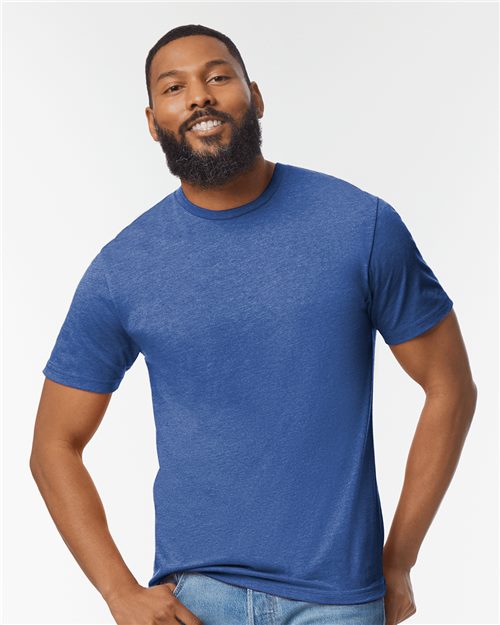 A bearded man smiles while wearing a blue Gildan Softstyle® Lightweight T-Shirt made from 100% ring-spun cotton and jeans, set against a plain, light background, representing socially conscious manufacturing.