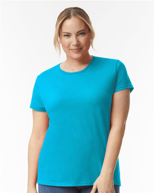 A person in a bright blue Gildan Softstyle® Women’s Lightweight T-Shirt made from ring-spun cotton stands against a plain white background, with light brown hair tied back, smiling slightly while looking directly at the camera.