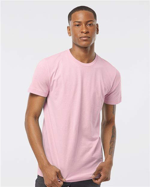 A person wears a Tultex Fine Jersey T-Shirt in plain pink, made from ring-spun cotton certified by OEKO-TEX Standard 100. They stand against a light gray backdrop, short-haired and looking forward, with one thumb casually hooked in a pocket.