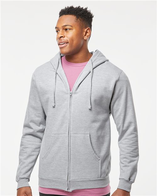 A person with short hair wears a Tultex light gray full-zip hooded sweatshirt made of soft ring-spun cotton over a pink shirt, standing against a plain white background and gazing slightly to the side with a neutral expression.