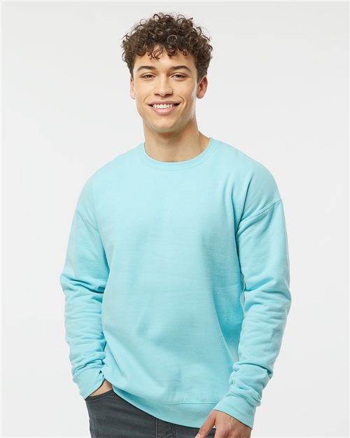 A person with curly hair smiles, wearing a light blue Tultex Fleece Crewneck Sweatshirt made from ring-spun cotton. Their hands rest in their pockets against a plain light gray background, emphasizing the sustainable comfort of the fabric.