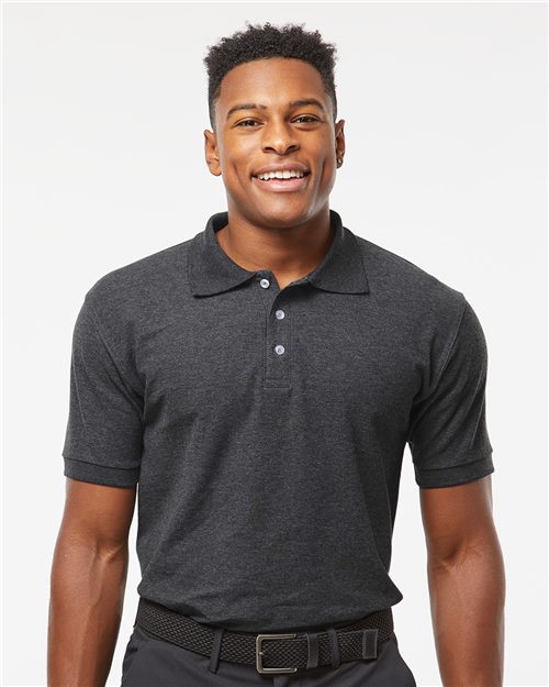 A person in a Tultex - 50/50 Sport Pique Polo in dark gray, made from a comfortable cotton/polyester blend, smiles against a white background. The shirt is buttoned up and paired with a black belt. The individual has short hair and an approachable demeanor.