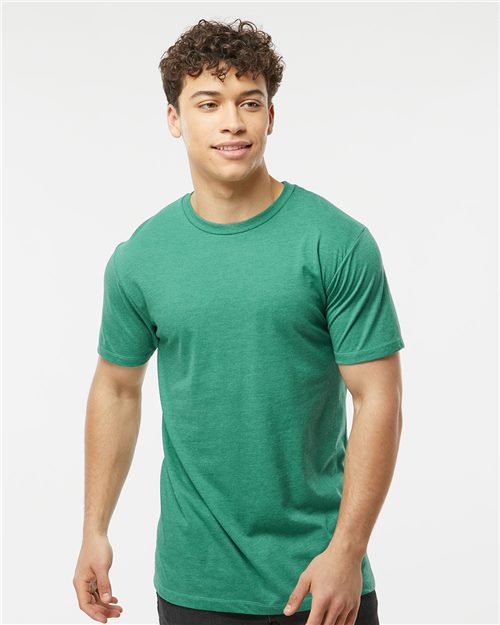 A curly-haired person is wearing a Tultex Premium Cotton Blend T-Shirt in green and black pants against a white background. They smile, glancing sideways, embodying effortless style and sustainable manufacturing principles.