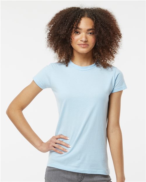 A person with curly hair, confidently wearing a Tultex Women's Fine Jersey Slim Fit T-Shirt in light blue and gray pants, poses with one hand on their hip and a slight smile, showcasing the spirit of sustainable manufacturing set against a plain white background.