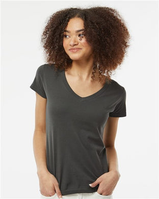 A curly-haired person wears a Tultex Women's Fine Jersey V-Neck T-Shirt, made from 100% ring-spun USA cotton, paired with light pants. They stand against a plain white background, slightly turned to the side and relaxed, representing sustainable manufacturing.