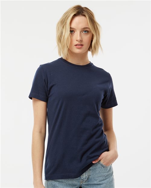 A person with short blonde hair, wearing a plain navy Tultex Women's Fine Jersey Classic Fit T-Shirt and light blue jeans, stands against a white background. They have their right hand in their pocket and glance slightly left.