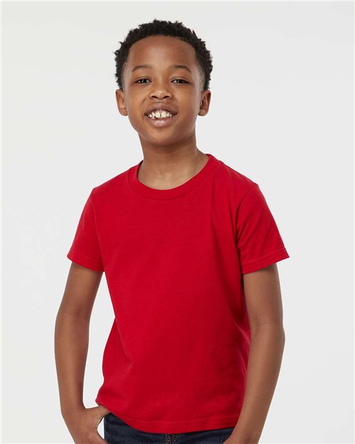 A smiling child wearing a Tultex - Youth Fine Jersey T-Shirt in bright red stands against a white background. The OEKO-TEX Standard 100-certified, ring-spun cotton shirt complements their dark jeans and short curly hair as their hands rest in their pockets.