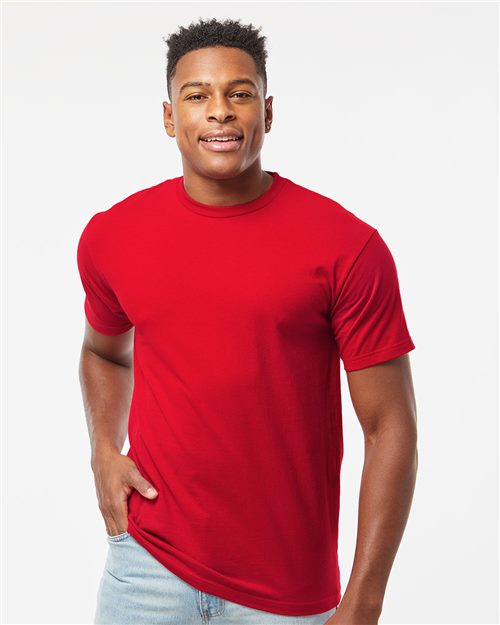 A person wearing a Tultex Heavyweight Jersey T-Shirt in bright red, made from USA cotton, and light blue jeans stands against a plain white background. They have short dark hair and are smiling with one hand in their pocket.