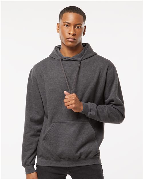 A person in a Tultex Fleece Hooded Sweatshirt, made from ring-spun cotton, stands against a plain white background. With one hand on the drawstring and wearing a neutral expression, this dark gray hoodie embodies the future of sustainable manufacturing with its eco-friendly design.