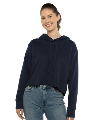 A woman wearing a Next Level Women's Laguna Sueded Raw Edge Crop Hoodie in navy blue and blue jeans smiles against a white background.