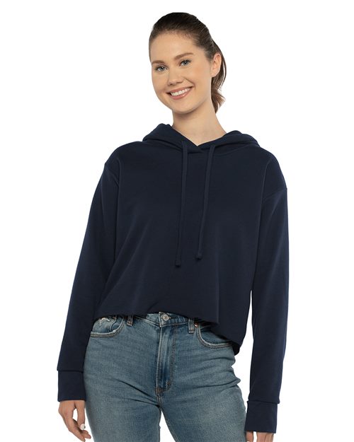 A woman wearing a Next Level Women's Laguna Sueded Raw Edge Crop Hoodie in navy blue and blue jeans smiles against a white background.