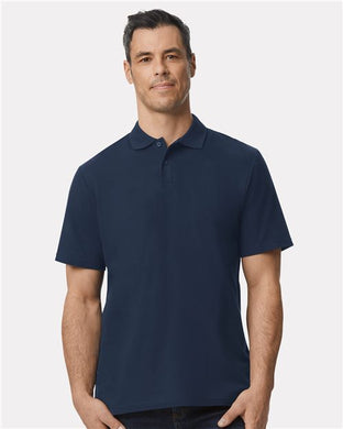 A man models the Gildan Softstyle® Pique Polo in dark blue against a plain white backdrop. His short dark hair and neutral expression enhance the scene's simplicity. The polo, known for its ring-spun cotton comfort, is a product of the Gildan brand.