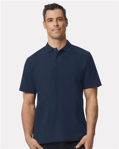 A man models the Gildan Softstyle® Pique Polo in dark blue against a plain white backdrop. His short dark hair and neutral expression enhance the scene's simplicity. The polo, known for its ring-spun cotton comfort, is a product of the Gildan brand.
