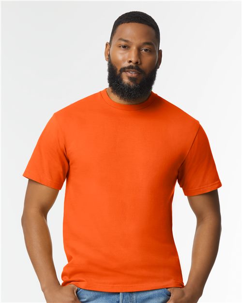 A bearded man stands against a plain white background, wearing a bright orange Gildan - Softstyle® Midweight T-Shirt and blue jeans. His hands are relaxed by his sides as he looks directly at the viewer.