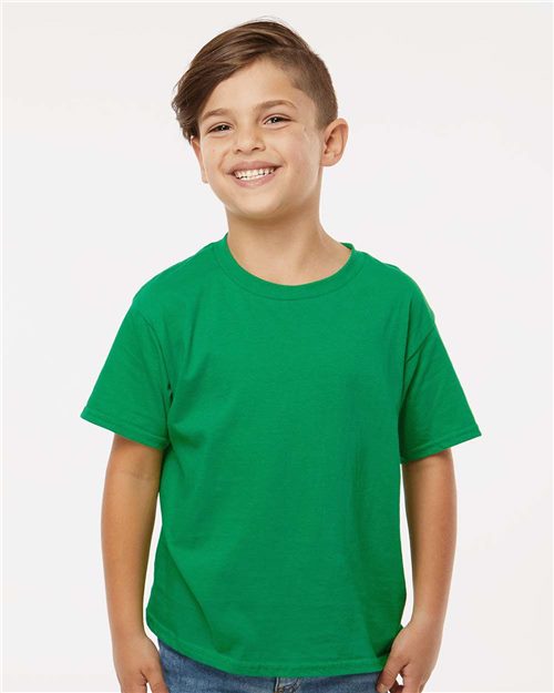 A smiling child with short, light brown hair wears a Gildan - Softstyle® Youth T-Shirt in green and blue jeans, standing against a plain white background.