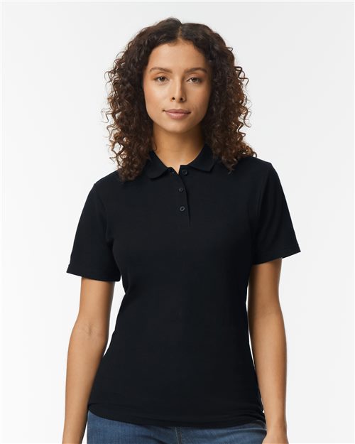 A woman with curly hair wears a Gildan Softstyle® Women's Pique Polo in black, made from OEKO-TEX certified ring-spun cotton. She stands against a white background, gazing directly at the camera, embodying sustainable manufacturing.