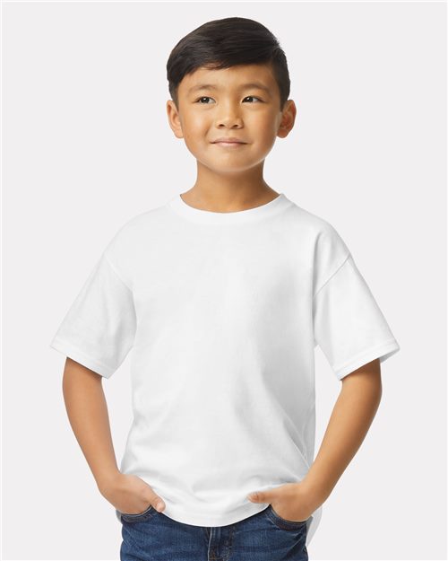 Load image into Gallery viewer, A young boy with short hair stands against a light background wearing a Gildan - Softstyle® Youth Midweight T-Shirt in plain white OEKO-TEX certified ring-spun cotton and blue jeans. He has his hands in his pockets and looks slightly upward with a neutral expression.
