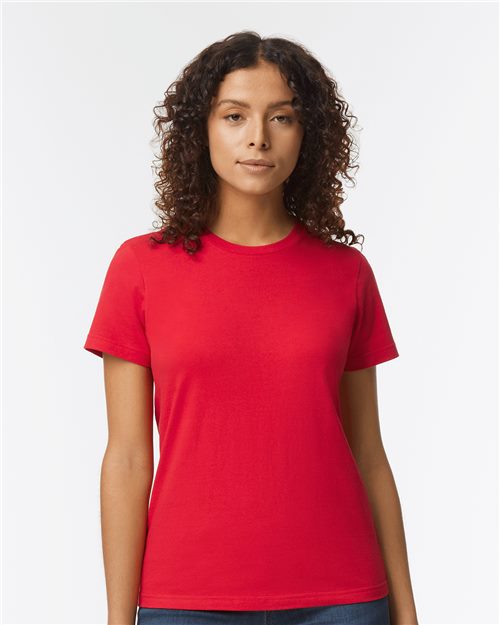 A curly-haired person wears a plain red Gildan Softstyle® Women's Midweight T-Shirt, made from OEKO-TEX certified ring-spun cotton, and jeans, standing against a white background while looking directly at the camera.