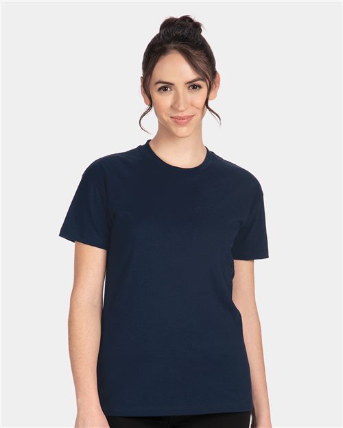 A person with dark hair tied up is wearing the Next Level - Women's Cotton Relaxed T-Shirt by Next Level, in plain navy blue, made from 100% combed ring-spun cotton. They stand against a light grey background, smiling slightly at the camera with their hands by their sides.