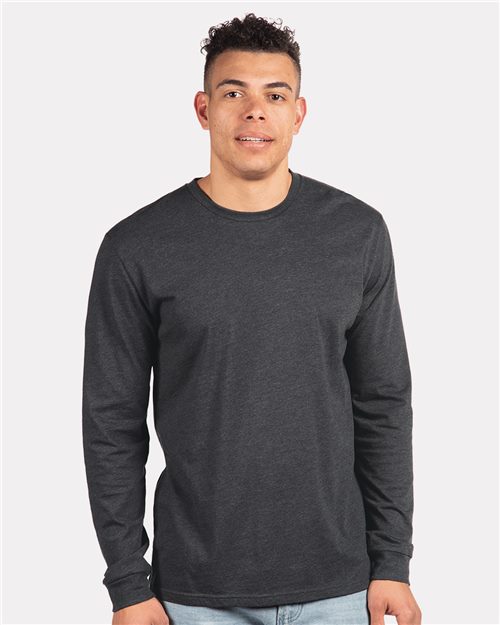 Wearing a Next Level CVC Long Sleeve T-Shirt in dark gray and light blue jeans, a person smiles gently at the camera against a plain light gray background.