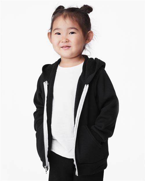 Load image into Gallery viewer, A young child with two small buns in their hair is smiling, wearing a BELLA + CANVAS Toddler Sponge Fleece Full-Zip Hoodie over an Airlume cotton BELLA + CANVAS shirt. Their hands rest in their pockets against a plain white background.
