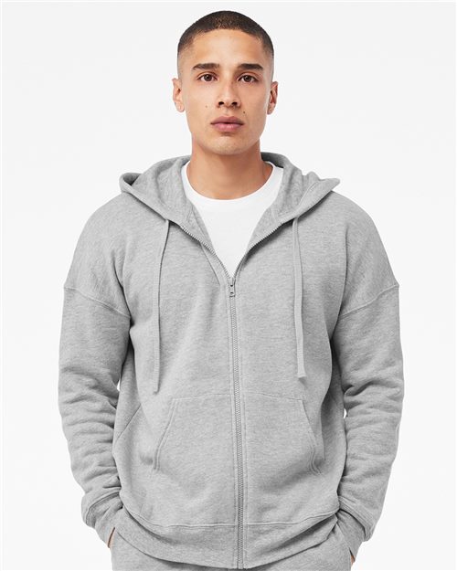 A person in a BELLA + CANVAS Sponge Fleece DTM Full-Zip Hoodie, made sustainably with Airlume combed cotton, stands against a white background. The grey hoodie is loose-fitting, has adjustable hood drawstrings, and features unisex sizing for versatility.