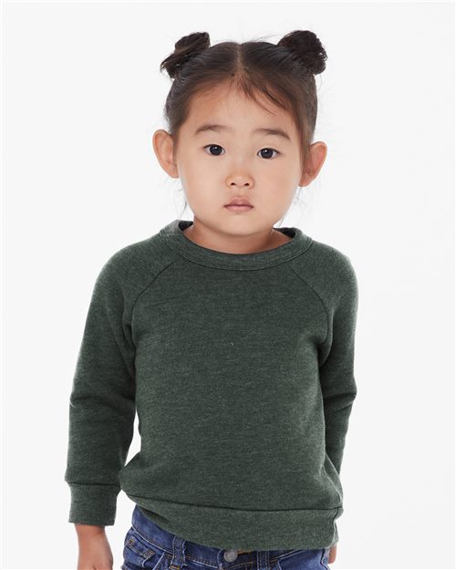 Load image into Gallery viewer, A child stands against a plain background in a BELLA + CANVAS Toddler Sponge Fleece Raglan Sweatshirt, made with Blue Sign certified dyes, and blue jeans. Their hair is styled in two small buns, highlighting the blend of style and eco-consciousness.
