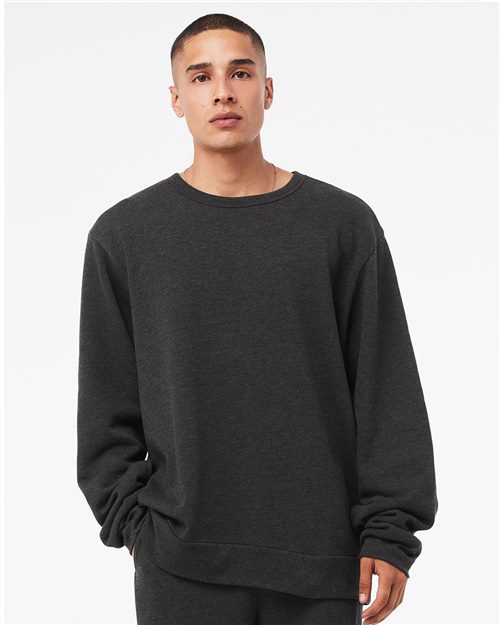 A person with short hair wears a loose-fitting, long-sleeved BELLA + CANVAS Sponge Fleece Classic Crewneck Sweatshirt, made from Airlume combed cotton in dark gray. Posed against a white background with one hand in their pocket, they highlight sustainable manufacturing and unisex sizing.