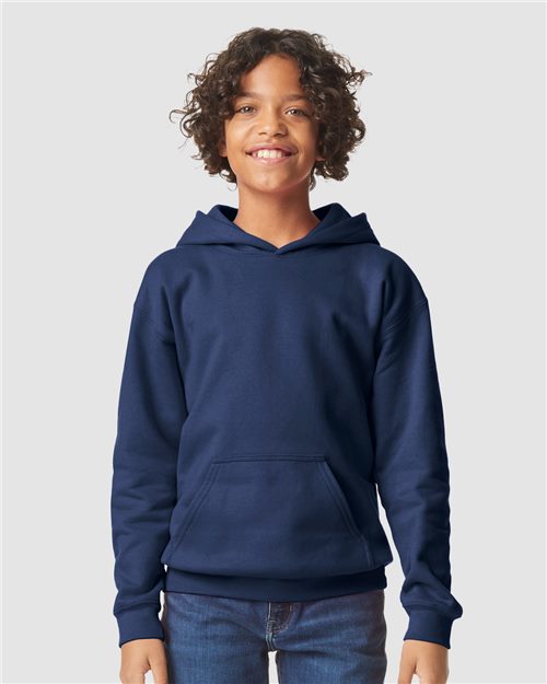 A smiling child with curly hair wears a Gildan Softstyle® Youth Midweight Hooded Sweatshirt in navy blue and jeans, set against a plain light background.