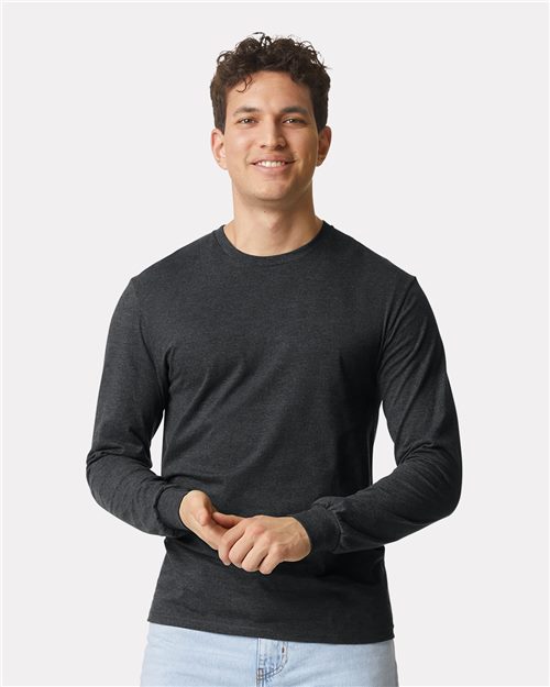 A smiling person in a Gildan Softstyle® CVC Long Sleeve T-Shirt (dark gray) and light blue jeans stands against a white background, showcasing the comfort of this modern classic fit made from premium ring-spun cotton.