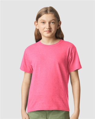 A person with long brown hair is wearing a bright pink Gildan - Softstyle® Youth CVC T-Shirt and olive green pants, standing against a plain gray background.